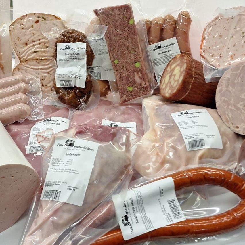 Eine Auswahl an verpackten Fleischsorten, einschließlich Würsten, Salami und Speck, ist auf einer weißen Oberfläche zu sehen. Jedes Fleisch hat einen Etikett mit Angaben wie Name, Gewicht und Zutaten.