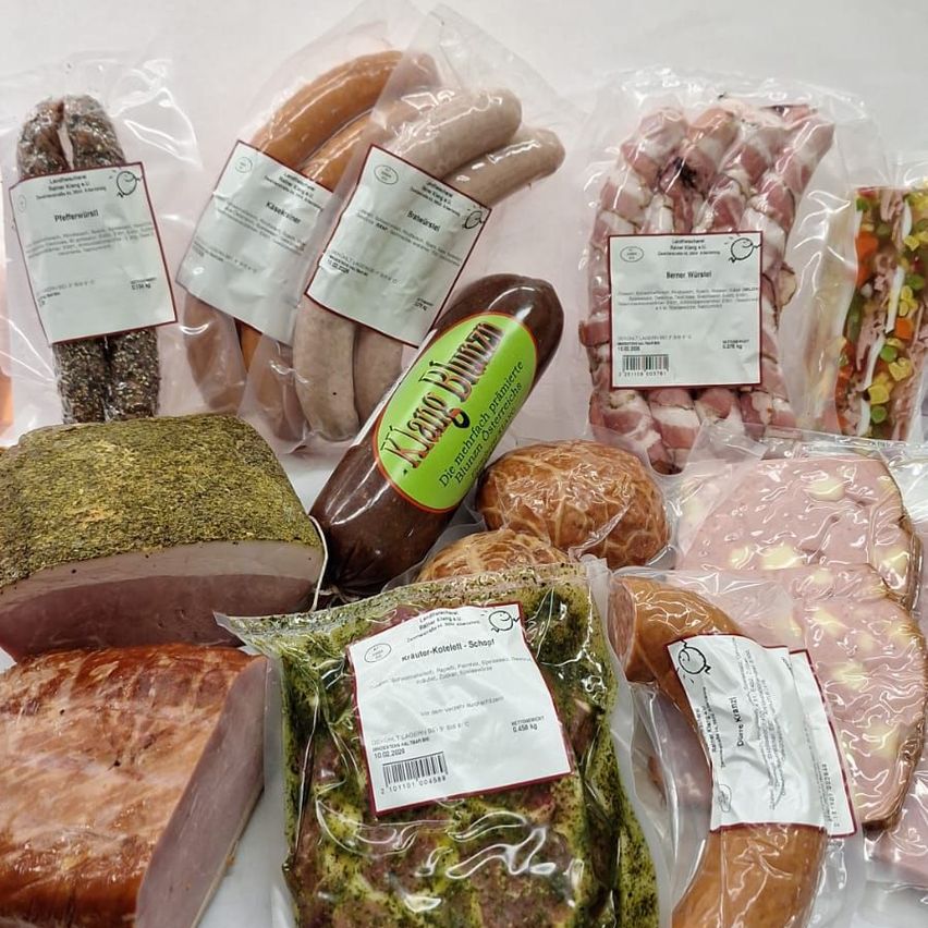 Eine Auswahl an Fleischsorten in Plastikverpackungen auf einer weißen Oberfläche, einschließlich Würste, Schinken, Speck und einige mit Etiketten. Die Fleischsorten umfassen 'Wurstwurst', 'Berzer Wursterl' und 'Krauter-Koteletts'. Einige sind geschnitten.