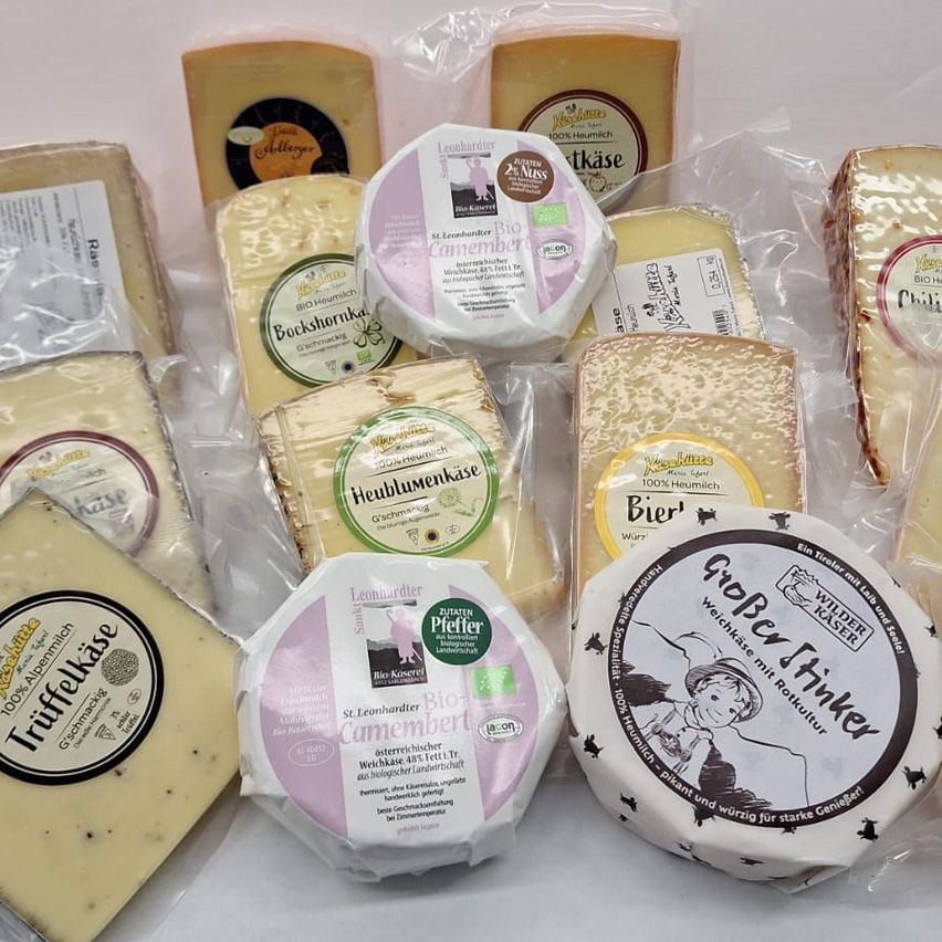 Eine Auswahl an Käsesorten, jede einzeln in Plastik verpackt. Jeder Käse hat ein Etikett, das seine Art, Herkunft und einige sind mit Bio-Zertifizierungszeichen gekennzeichnet. Es gibt mehrere Sorten, darunter Camembert, Heublumenkäse und andere.