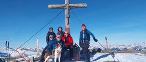 Eine Gruppe von Menschen mit einem Hund posiert für ein Foto auf einem verschneiten Berggipfel vor einem großen Holzkreuz.