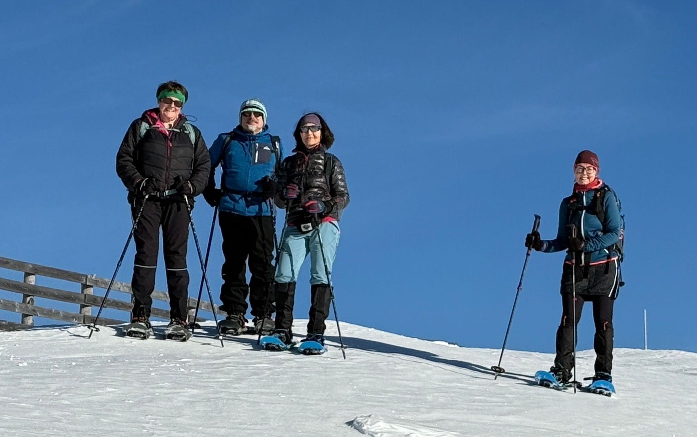 Vier Personen, alle für Wintersport gekleidet, stehen auf einem verschneiten Hang und lächeln für ein Foto. Jeder ist mit Skistöcken und Skischuhen ausgestattet.