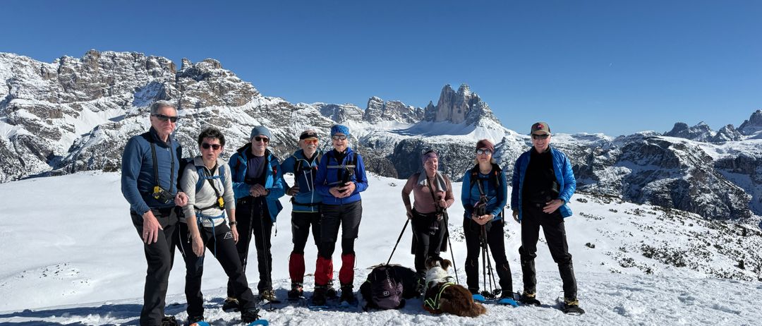 Eine Gruppe von Wanderern in Winterkleidung posiert für ein Foto im Schnee mit einem Hund davor. Schneebedeckte Berge und klarer blauer Himmel im Hintergrund.