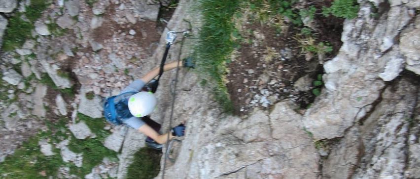 Eine Person mit Helm und Gurt klettert einen felsigen Berghang hinauf, hält eine Seil und benutzt einen Pickel.