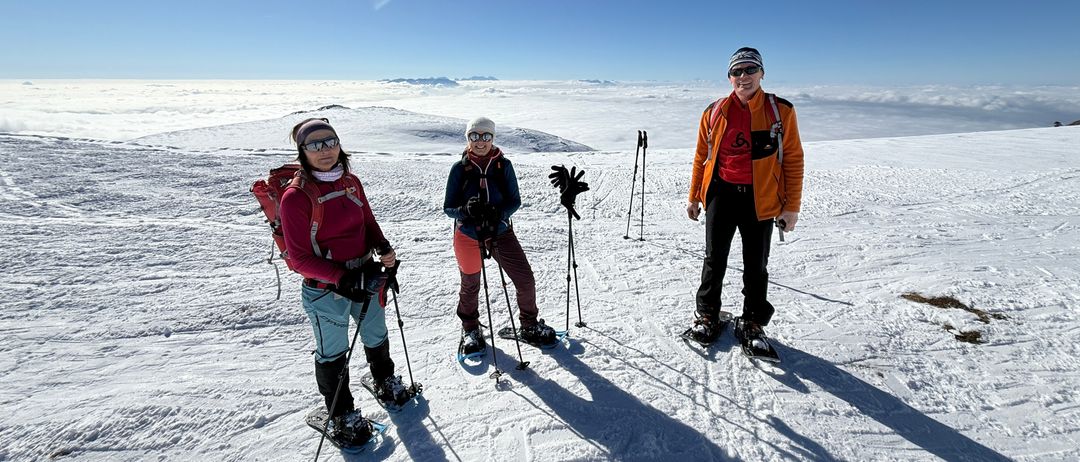 Drei Personen, ausgestattet mit Winterkleidung und Schneeschuhen, stehen auf einem verschneiten Berg unter einem blauen Himmel. Sie lächeln und posieren für ein Foto.