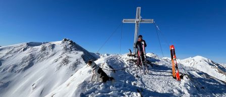 Ein Skifahrer steht auf einem verschneiten Berg, in der Nähe eines Kreuzes, mit einem Hund an der Leine. Der Himmel ist klar und blau.