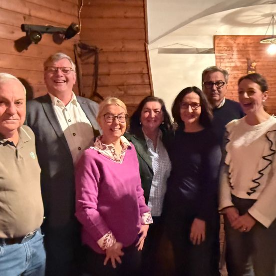 Sieben Personen stehen in einem Raum mit Holzwänden, eine Frau trägt einen lila Pullover und eine Brille und sie alle lächeln.