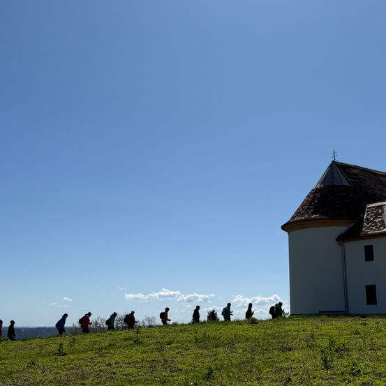 Eine Gruppe von Wanderern geht einen grasbewachsenen Hügel hinauf, im Hintergrund ein Gebäude.