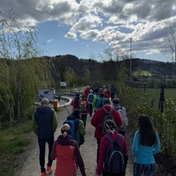 Eine Gruppe von Menschen mit Rucksäcken und Turnschuhen geht auf einem Kiesweg. Der Weg ist von Grünflächen und einem Zaun umgeben. Hinter dem Zaun befindet sich eine Rasenfläche mit Bäumen und Büschen.
