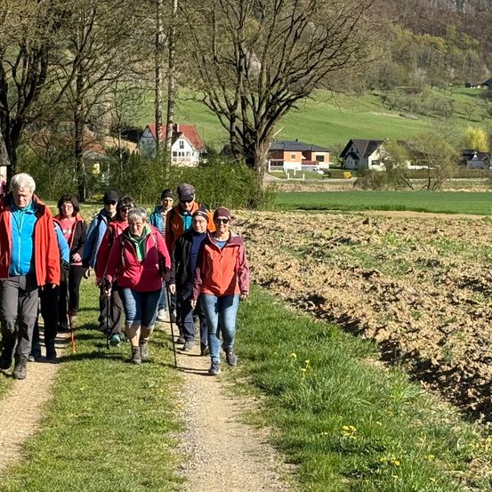 Eine Gruppe von Wanderern geht auf einem Grasweg in einem ländlichen Gebiet. Einige tragen Hüte und Brillen, und einige haben Stöcke. Im Hintergrund gibt es Häuser, Bäume und ein Grasfeld.