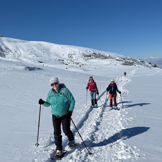 Drei Frauen, die auf einem verschneiten Berg schneeschuhwandern. Sie tragen Winterkleidung und haben Skistöcke dabei.