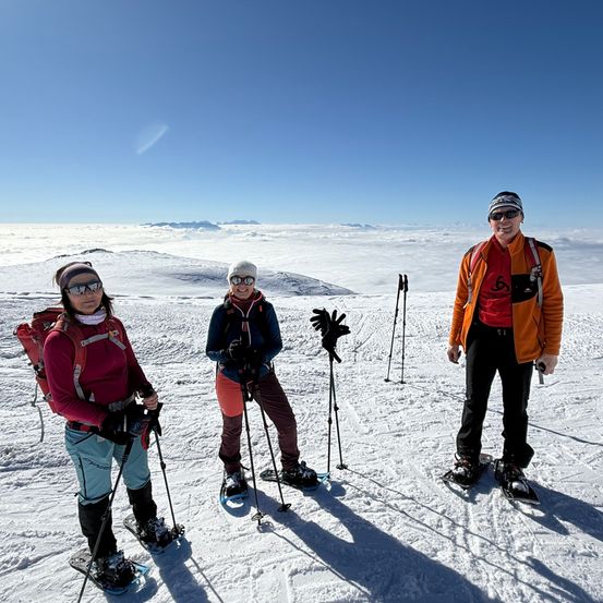 Drei Personen posieren für ein Foto auf einem verschneiten Berg mit Rucksäcken und Schneeschuhen.
