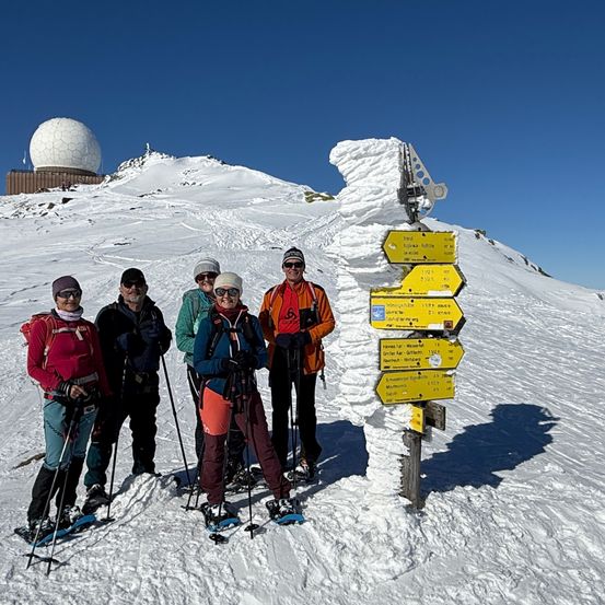 Eine Gruppe von Menschen steht im Schnee vor einem Schild, trägt Skier und Skistöcke. Hinter ihnen befindet sich ein verschneiter Berg mit einem Gebäude und einer Radarkuppel oben.