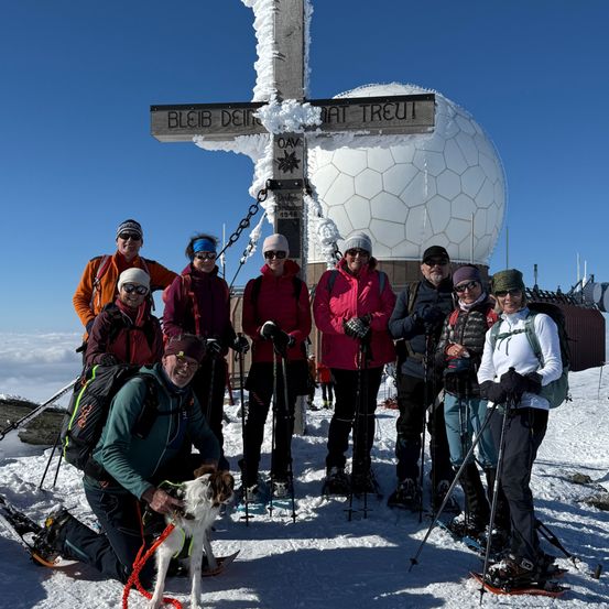 Eine Gruppe von Menschen in Winterkleidung und mit Skistöcken posiert für ein Foto vor einem Schild und einer Radarkuppel auf einem verschneiten Berg.