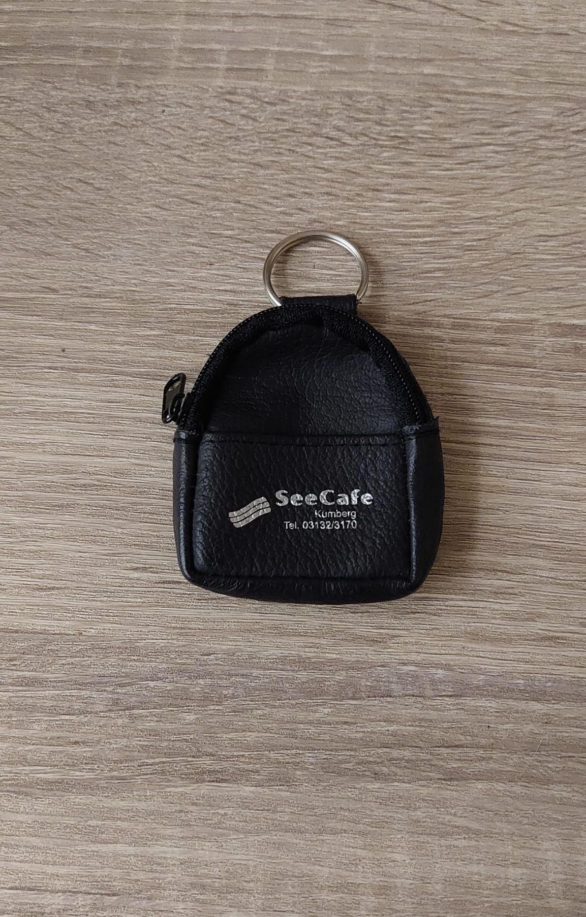 Ein schwarzer Lederschlüsselanhänger mit Reißverschluss, der das SeeCafe-Logo und Kontaktinformationen trägt, liegt auf einer Holzoberfläche.