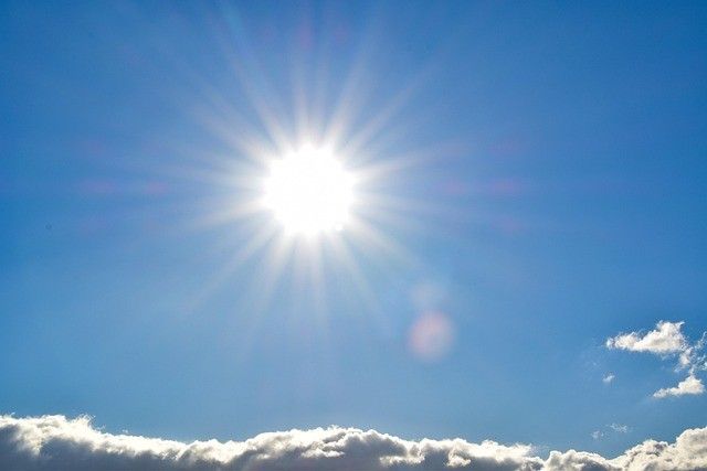 A bright sun shines through a clear blue sky with a few scattered clouds.