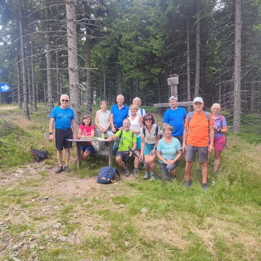Eine Gruppe von Wanderern in Freizeitkleidung posiert für ein Foto im Wald, mit einem Holztisch und einem Kreuz im Hintergrund.