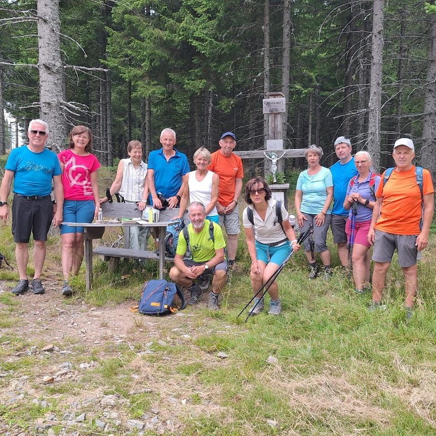 A group of hikers, dressed in casual outdoor clothing, poses for a photo in a forest clearing with a wooden cross and bench in the background.
