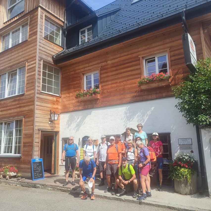 A group of hikers in front of a wooden building with a blue roof, some wearing hats and carrying backpacks, posing for a photo.