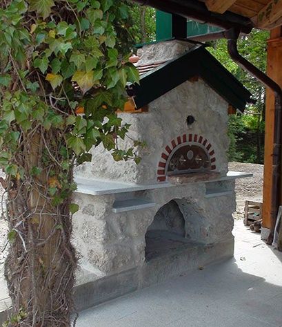 Ein Steinbackofen im Freien mit schwarzem Dach und roter Einfassung, umgeben von Grün und Bäumen, mit einem Holzpfosten links.