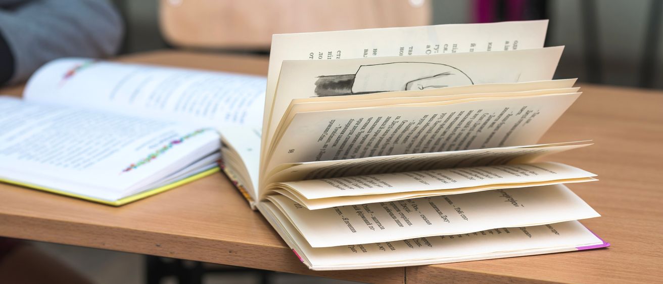 Ein offenes Buch liegt auf einem Schreibtisch, mit ausgefächerten Seiten. Ein Holzstuhl ist im Hintergrund. Ein weiteres Buch und ein Notizbuch sind teilweise auf der linken Seite zu sehen.