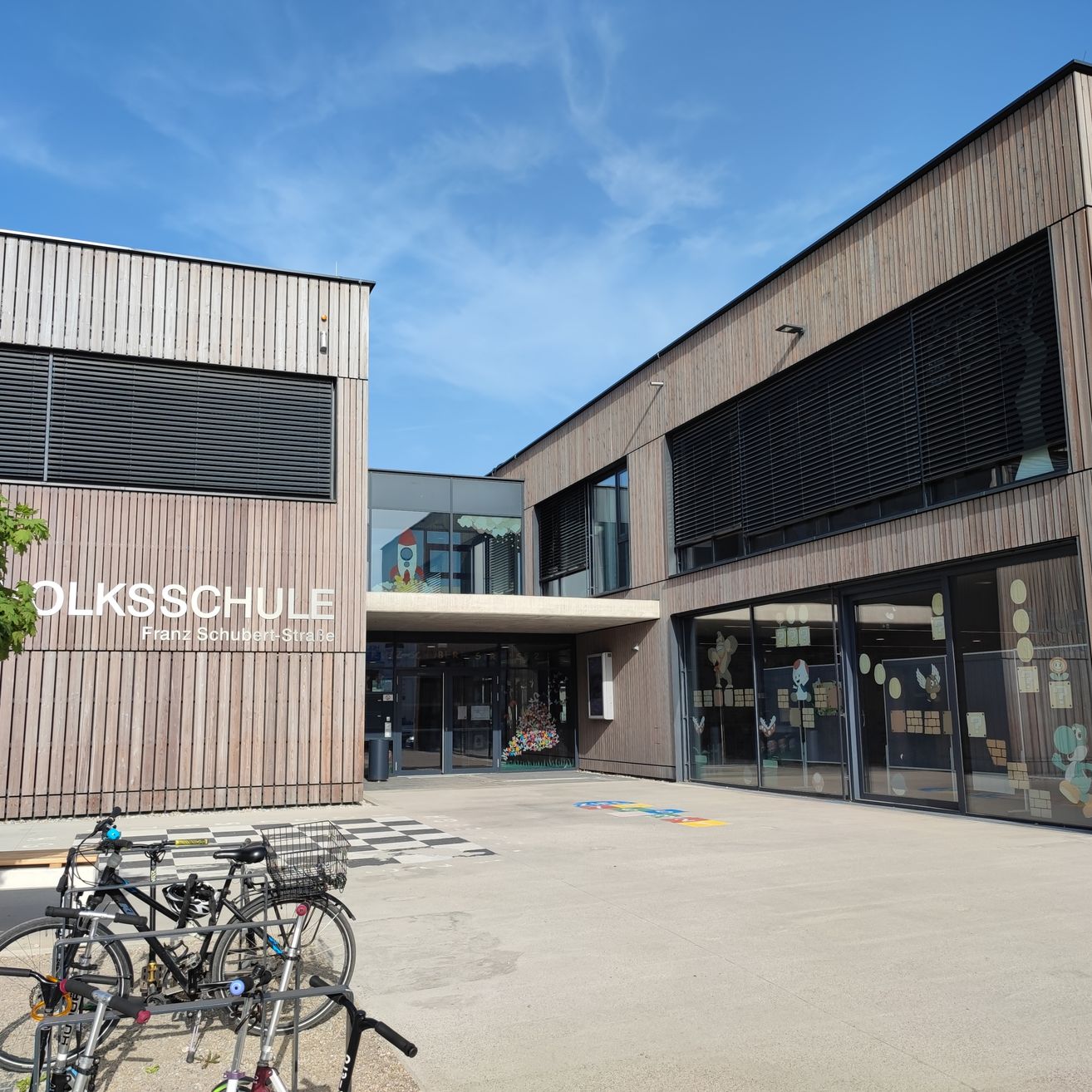 Ein Gebäude mit dem Wort 'OLKSSCHULE' und 'Franz Schubert Straße' darauf. Fahrräder sind davor geparkt.