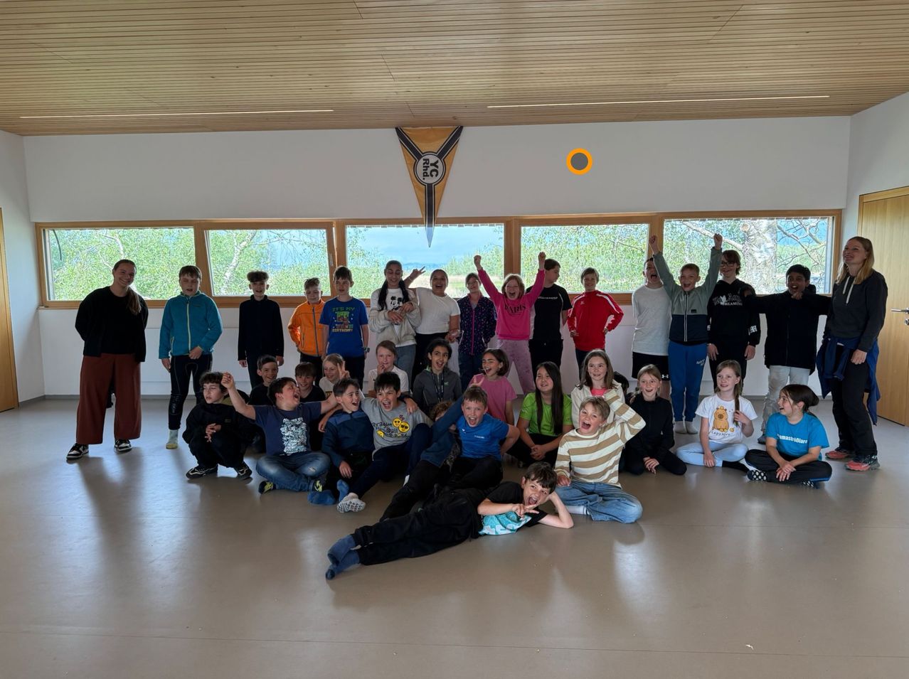 Eine Gruppe junger Menschen posiert für ein Foto in einem Raum mit Holzdecke, lächelt und macht Gesten. Einige stehen, andere sitzen auf dem Boden. Es gibt ein Banner mit Text und Symbol.