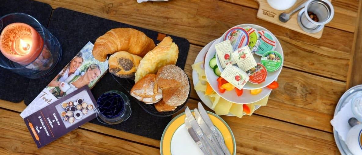 Ein Holztisch mit verschiedenen Frühstücksartikeln. Teller halten Croissants, Brot und Käse. Tassen mit Kaffee und Tee sind mit Untertellern bestückt. Eine Kerze und ein Messerset sind ebenfalls auf dem Tisch.