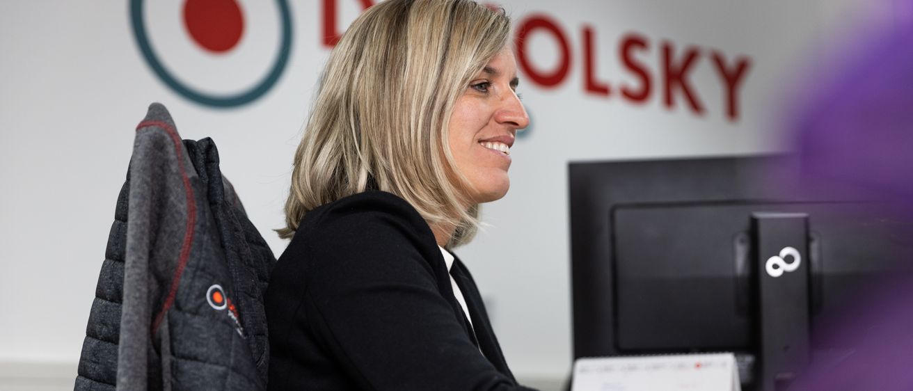 Eine Frau mit blonden Haaren lächelt, während sie an einem Schreibtisch vor einem Computer sitzt. Sie trägt ein schwarzes Oberteil und sitzt hinter einer Wand mit einem Logo.