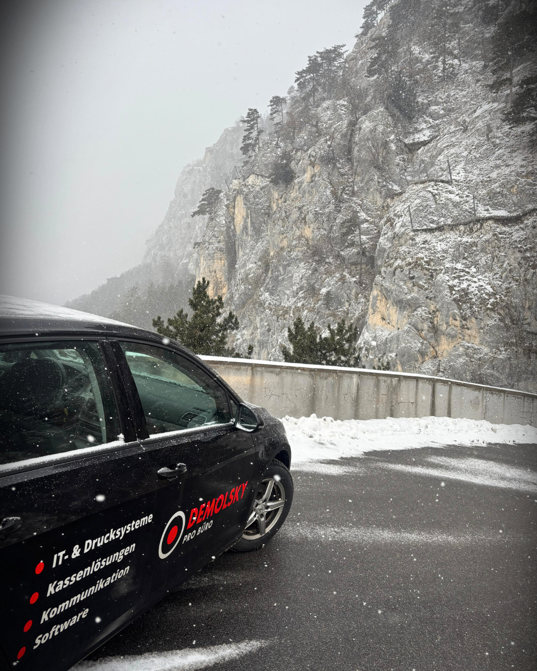A black car is parked on a snowy road with a mountain covered in snow in the background. The car has a logo and text on its side that reads 'IT & Drucksysteme Kassenlösungen'.