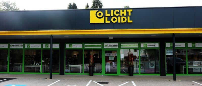 Das Äußere eines Geschäfts namens O Licht Loidl, mit einem schwarzen und gelben Dach, grünen Fenstern und zwei Topfpflanzen. Das Geschäft hat Glastüren und einen Parkplatz mit weißen Markierungen.