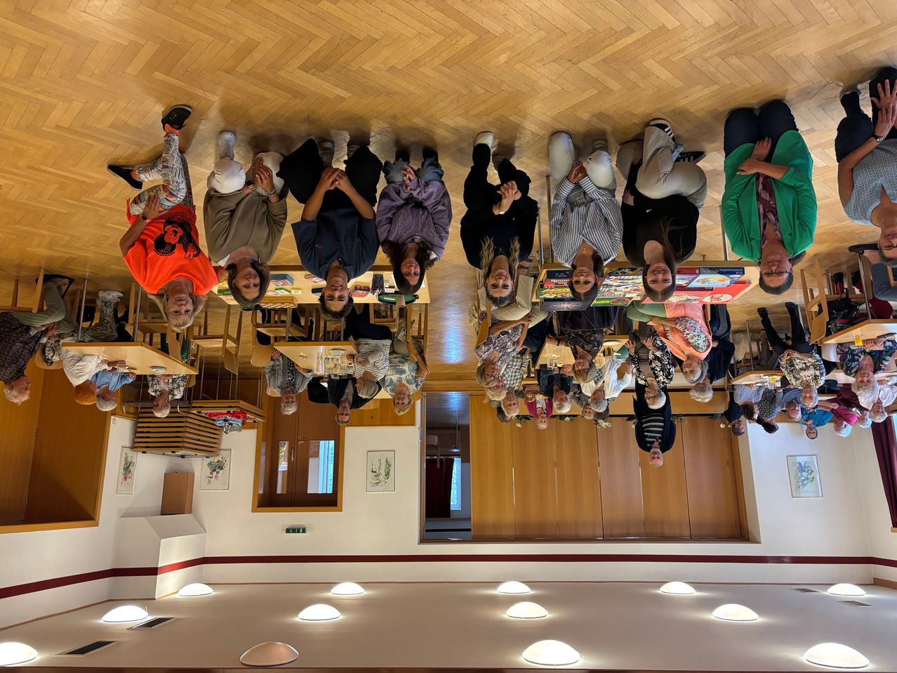 Eine Gruppe von Menschen steht in einem Kreis in einem Raum, lächelt und posiert für ein Foto. Sie tragen Freizeitkleidung. Der Raum hat Holzböden und Wände, mit mehreren Tischen und Stühlen. Es hängen Bilderrahmen an der Wand, und eine Tür befindet sich hinten.