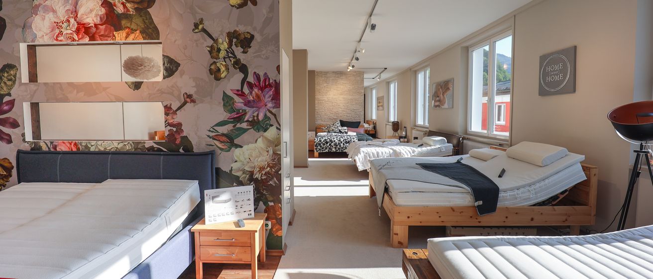 Das Innere eines Schlafzimmers mit Holzbetten auf weißem Teppich. Eine Wand ist mit einem großen Tapetenmuster mit Blumendesigns verziert. Ein hölzerner Nachttisch steht auf der linken Seite.