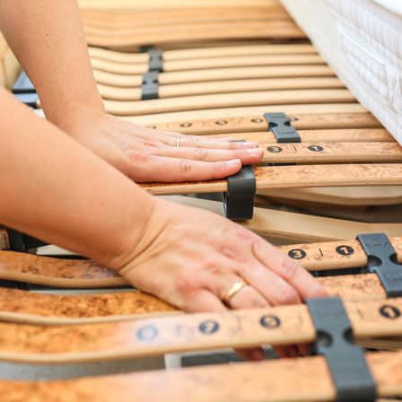 Eine Person justiert die Lamellen eines nummerierten Holzbettrahmens, ausgestattet mit schwarzen Clips, während sie einen Ring am Finger trägt.