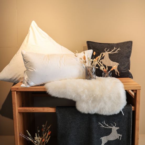 Ein Holztisch zeigt drei Kissen und eine Felldecke. Ein dekoratives Kissen mit Hirsch-Print liegt oben auf den Kissen. Ein Blumenstrauß aus getrockneten Blumen steht neben dem Hirsch-Kissen.