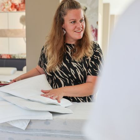 Eine Frau mit blonden Haaren, die ein schwarz-weiß gemustertes Shirt trägt, lächelt und hält ein weißes Kissen. Sie steht vor einem Tisch mit mehreren weißen Kissen. Im Hintergrund ist ein verschwommener Hintergrund mit einer weißen Wand und einem Regal mit einigen Gegenständen zu sehen.