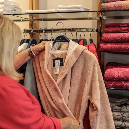 Eine Frau betrachtet einen pfirsichfarbenen Bademantel in einem Bekleidungsgeschäft, mit roten Handtüchern auf den Regalen hinter ihr.
