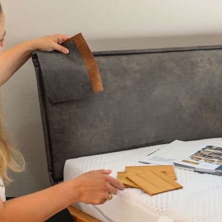 Eine Frau justiert die graue Polsterung eines Kopfteils. Sie hält ein Stück braunes Material, wahrscheinlich zur Reparatur oder Anpassung. Zusätzliches Material liegt auf dem Bett.