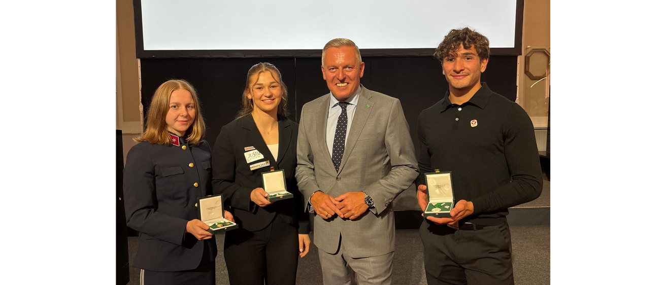Drei Personen posieren auf einem roten Teppich mit Auszeichnungen in der Hand, ein Mann in Anzug steht zwischen zwei Frauen in formeller Kleidung. Hinter ihnen ist eine große Leinwand, die das Logo von 'Das Land Steiermark' zeigt.