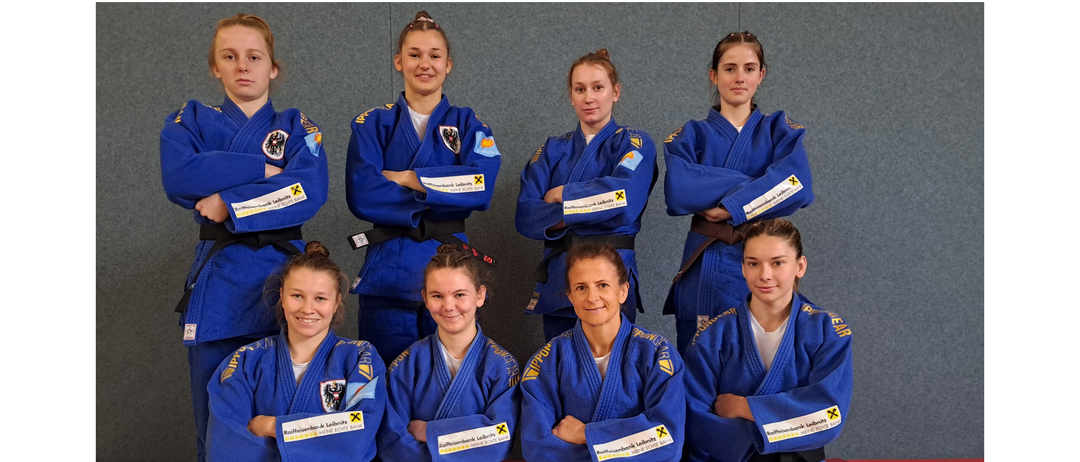 Eine Gruppe junger Frauen in blauen Kampfsportuniformen posiert für ein Foto auf einem roten Matten. Sie haben die Arme vor einer grauen Wand verschränkt.
