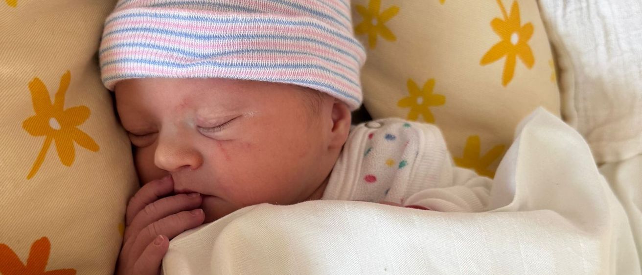 Ein Baby schläft friedlich, eingebettet in einem Kinderbett mit einem blumengemusterten Kissen. Das Baby trägt eine gestreifte Mütze und ist in eine weiße Decke gewickelt.