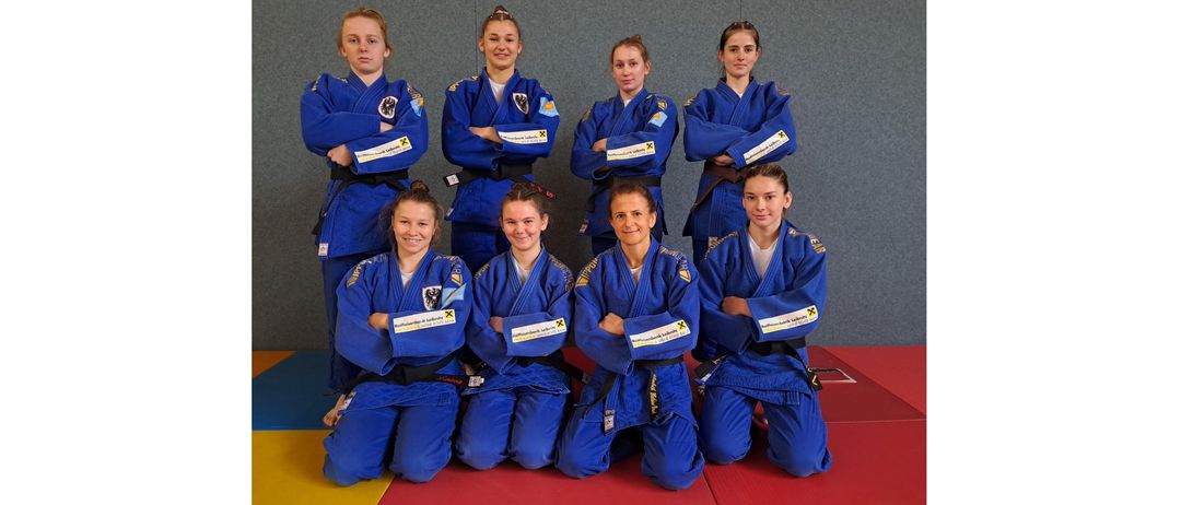 Eine Gruppe junger Frauen in blauen Kampfsportuniformen posiert für ein Foto. Sie alle lächeln und haben die Arme verschränkt. Sie tragen schwarze Gürtel.