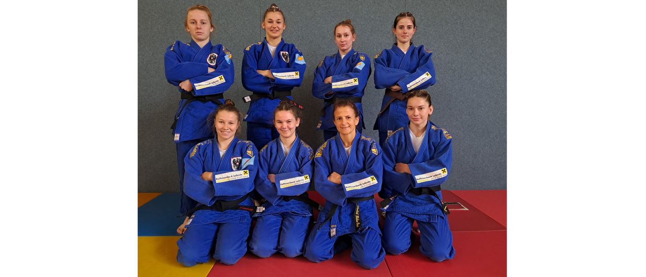 Eine Gruppe junger Frauen in blauen Kampfsportuniformen posiert für ein Foto. Sie alle lächeln und haben die Arme verschränkt. Sie tragen schwarze Gürtel.
