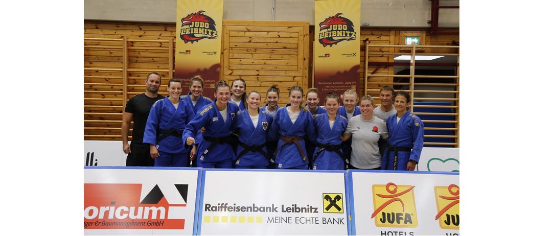 Eine Gruppe von Judoka steht in einer Turnhalle mit zwei Bannern hinter ihnen. Sie tragen blaue Uniformen. Ein Banner sagt 'JUDOLEIBNITZ' und das andere hat einen roten Drachen.