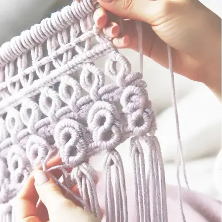 Eine Person hält ein graues gewebtes Gewebe mit einem Quastendesign, möglicherweise häkelnd oder strickend. Das Gewebe hat ein wellenförmiges Muster und Fransen an den Rändern.