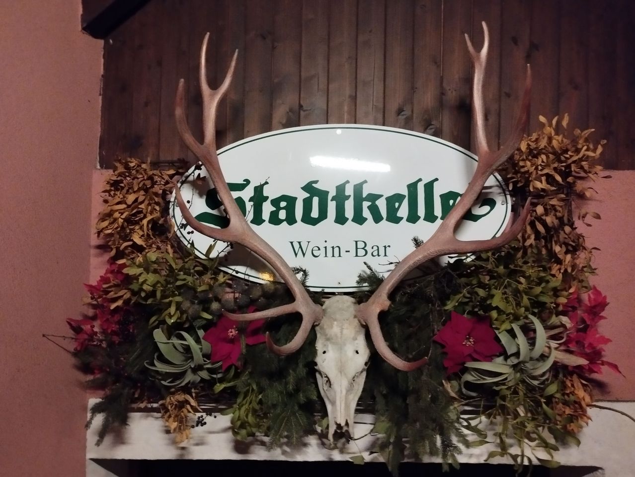 Eine Holzwand mit einem Schild, das Stadtkelle Wein-Bar besagt. Es ist mit einem Hirschschädel, Geweih und roten Blumen geschmückt.