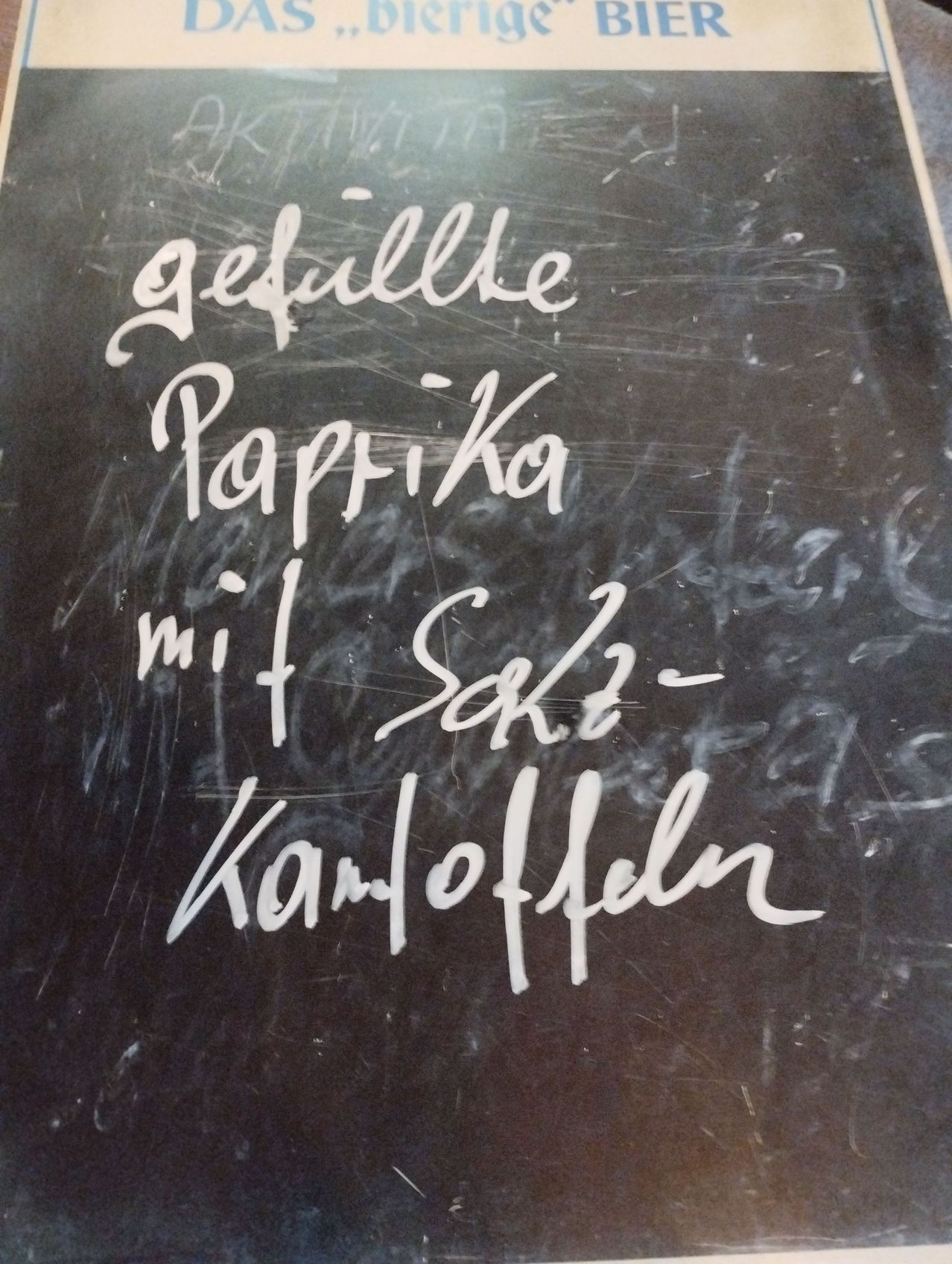 A blackboard with handwritten text in white chalk. It reads 'Gefüllte Paprika mit Salz-Kartoffeln'. The writing is somewhat faded and there are smudges on the board.