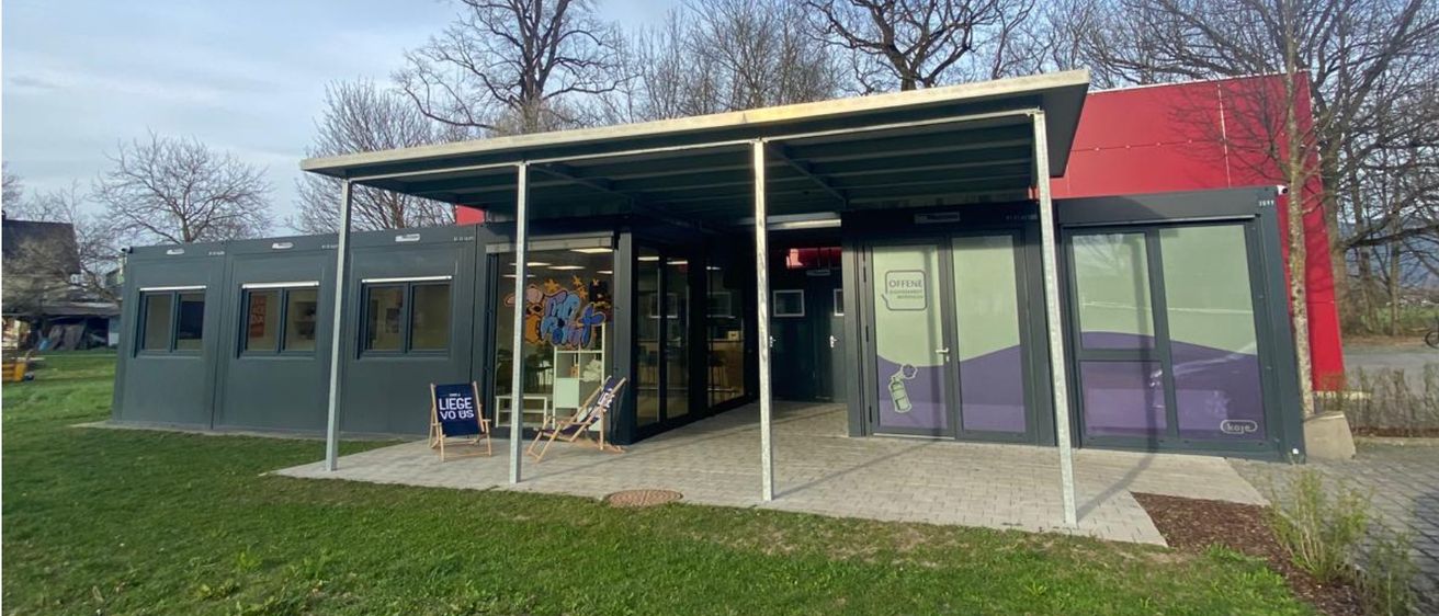 Ein modernes Modulgebäude mit Vordach steht auf einer Rasenfläche unter einem teilweise bewölkten Himmel. Das Gebäude hat Glastüren und Fenster, mit einem roten Gebäude im Hintergrund.