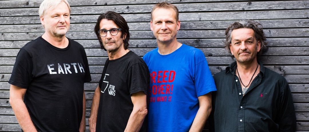 Vier Männer stehen vor einer Holzwand. Der Mann auf der linken Seite trägt ein schwarzes T-Shirt mit dem Wort Earth darauf. Der zweite Mann trägt eine Brille und ein schwarzes T-Shirt. Der dritte Mann trägt ein blaues T-Shirt mit der Aufschrift Reed Over. Der vierte Mann trägt ein schwarzes T-Shirt mit einer Halskette.