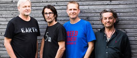 Vier Männer stehen vor einer Holzwand. Der Mann auf der linken Seite trägt ein schwarzes T-Shirt mit dem Wort Earth darauf. Der zweite Mann trägt eine Brille und ein schwarzes T-Shirt. Der dritte Mann trägt ein blaues T-Shirt mit der Aufschrift Reed Over. Der vierte Mann trägt ein schwarzes T-Shirt mit einer Halskette.