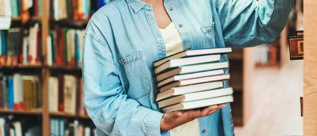Eine Frau hält einen Stapel Bücher in einer Bibliothek. Sie trägt eine Jeansjacke und ein weißes Unterhemd.